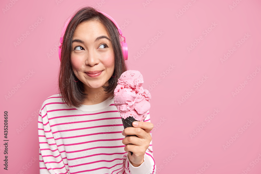 Studio shot of pleasant looking young Asian woman with dark hair holds big strawberry  ice cream looks away thoughtfully wears stereo headphones isolated over  pink background copy space area. Stock Photo
