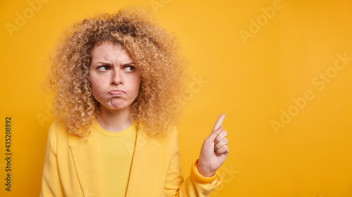 Curly haired angry woman looks with judgement points finger right stands upset blows cheeks makes irritated grimace dressed in formal outfit isolated over yellow background complains about something © wayhome.studio 