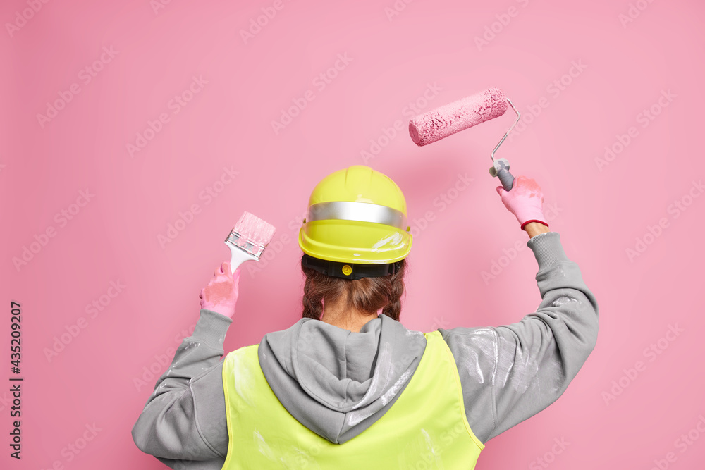 Faceless professional builder busy doing reconstruction of building stands back to camera uses equipment for painting walls wears protective hardhat and uniform poses against pink background Faceless professional builder busy doing reconstruction of building stands back to camera uses equipment for painting walls wears protective hardhat and uniform poses against pink background