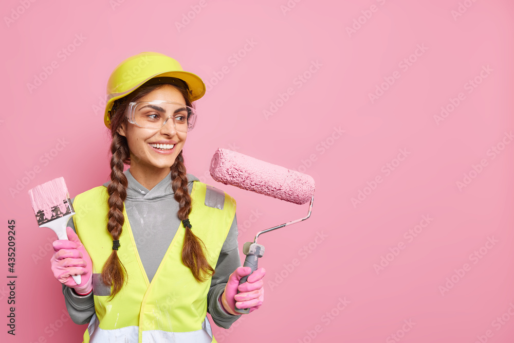 Positive busy female builder involved in renovation and repair of building holds roller and paint brush follows strict safety regulations wears protective equipment has leadership skills. Copy space Positive busy female builder involved in renovation and repair of building holds roller and paint brush follows strict safety regulations wears protective equipment has leadership skills. Copy space