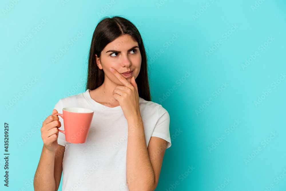 Young caucasian woman holding a mug isolated on blue background looking sideways with doubtful and skeptical expression. Young caucasian woman holding a mug isolated on blue background looking sideways with doubtful and skeptical expression.