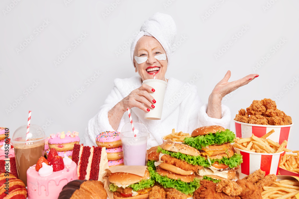 Positive carefree old woman wears shower towel on head beauty patches under eyes red lipstick cares about herself drinks soft drink eats junk food has unhealthy nutrition no restrictions and diets Positive carefree old woman wears shower towel on head beauty patches under eyes red lipstick cares about herself drinks soft drink eats junk food has unhealthy nutrition no restrictions and diets
