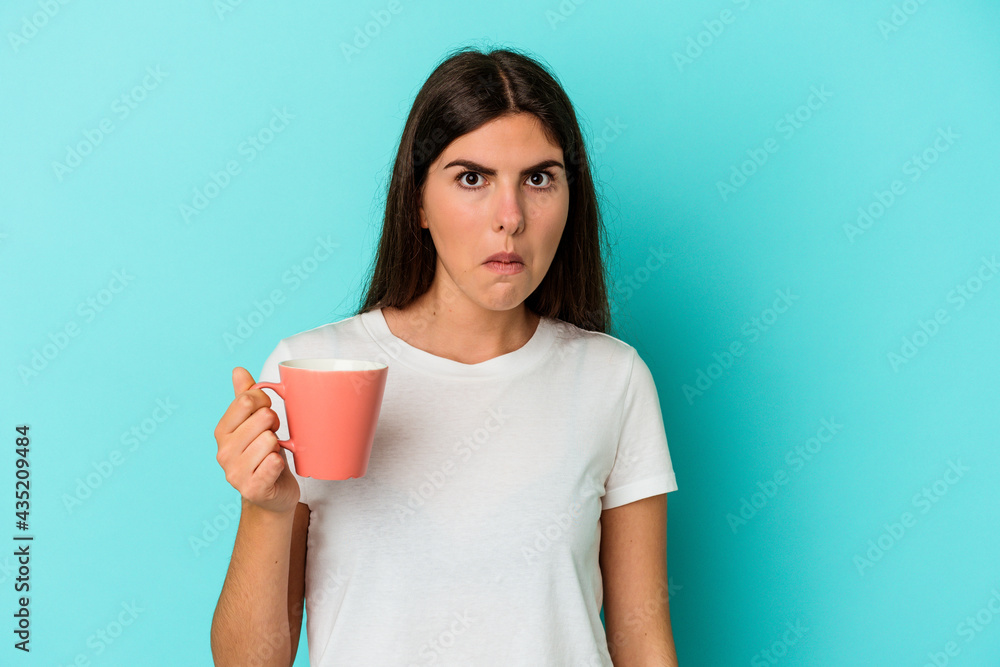 Young caucasian woman holding a mug isolated on blue background shrugs shoulders and open eyes confused. Young caucasian woman holding a mug isolated on blue background shrugs shoulders and open eyes confused.