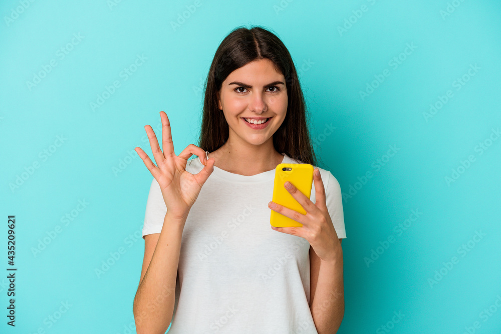 Young caucasian woman holding a mobile phone isolated on blue background cheerful and confident showing ok gesture.