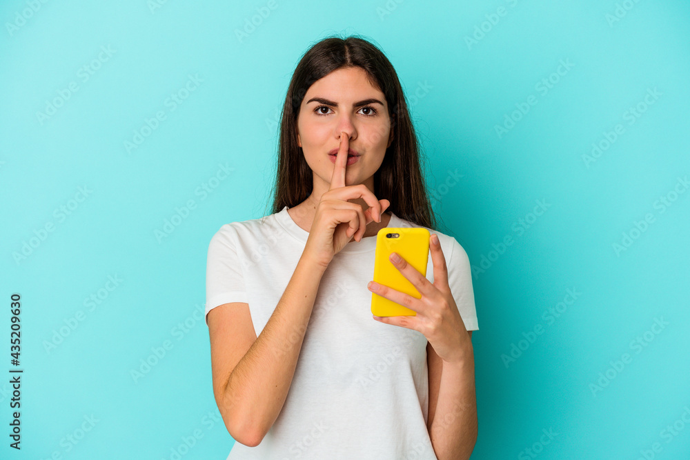 Young caucasian woman holding a mobile phone isolated on blue background keeping a secret or asking for silence. Young caucasian woman holding a mobile phone isolated on blue background keeping a secret or asking for silence.
