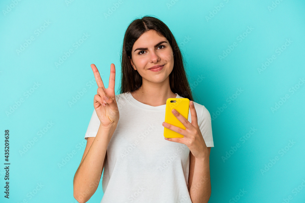 Young caucasian woman holding a mobile phone isolated on blue background showing number two with fingers. Young caucasian woman holding a mobile phone isolated on blue background showing number two with fingers.