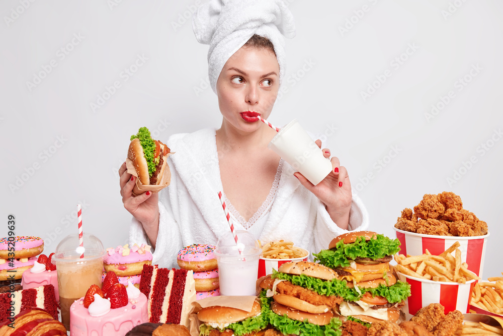 Thoughtful woman with clean fresh healthy skin dressed in bath robe wrapped towel on head drinks fizzy drink eats burger cannot refuse eating cheat meal surrounded by fast food stands indoor Thoughtful woman with clean fresh healthy skin dressed in bath robe wrapped towel on head drinks fizzy drink eats burger cannot refuse eating cheat meal surrounded by fast food stands indoor
