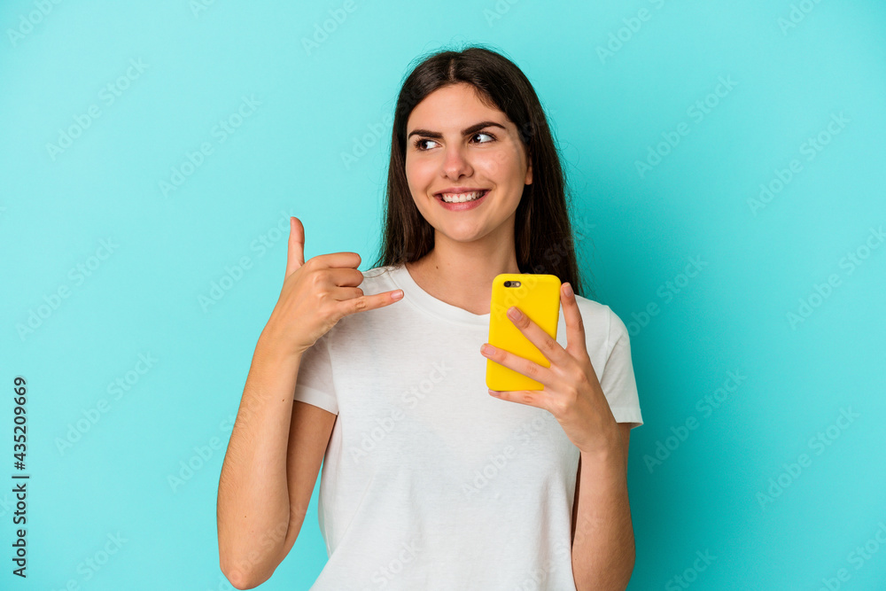 Young caucasian woman holding a mobile phone isolated on blue background showing a mobile phone call gesture with fingers. Young caucasian woman holding a mobile phone isolated on blue background showing a mobile phone call gesture with fingers.