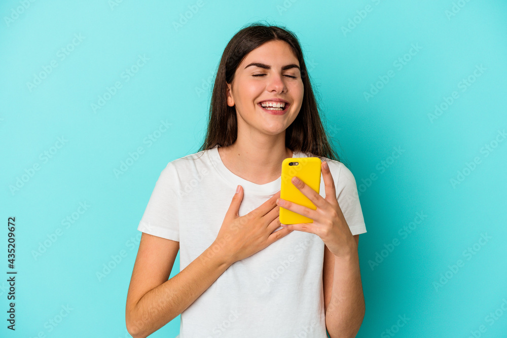 Young caucasian woman holding a mobile phone isolated on blue background laughs out loudly keeping hand on chest. Young caucasian woman holding a mobile phone isolated on blue background laughs out loudly keeping hand on chest.