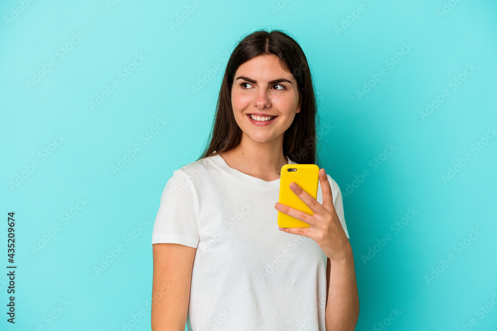 Young caucasian woman holding a mobile phone isolated on blue background looks aside smiling, cheerful and pleasant. Young caucasian woman holding a mobile phone isolated on blue background looks aside smiling, cheerful and pleasant.