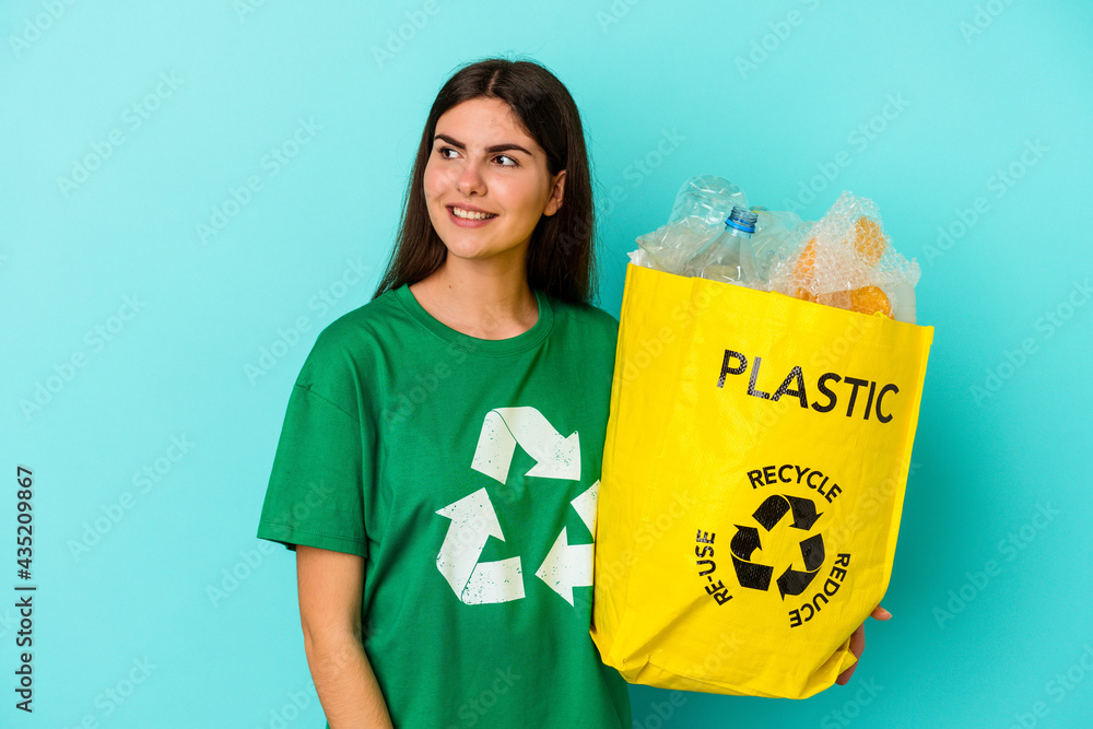 Young caucasian woman recycled plastic isolated on blue background looks aside smiling, cheerful and pleasant. Young caucasian woman recycled plastic isolated on blue background looks aside smiling, cheerful and pleasant.