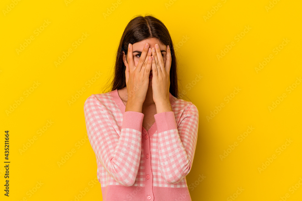 Young caucasian woman isolated on yellow background blink at the camera through fingers, embarrassed covering face. Young caucasian woman isolated on yellow background blink at the camera through fingers, embarrassed covering face.