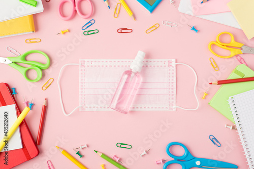 Top view photo of multicolor stationery scissors pens pencils pushpins clips calculator copybooks and sanitizer bottle on medical facemask in the middle isolated pastel pink background