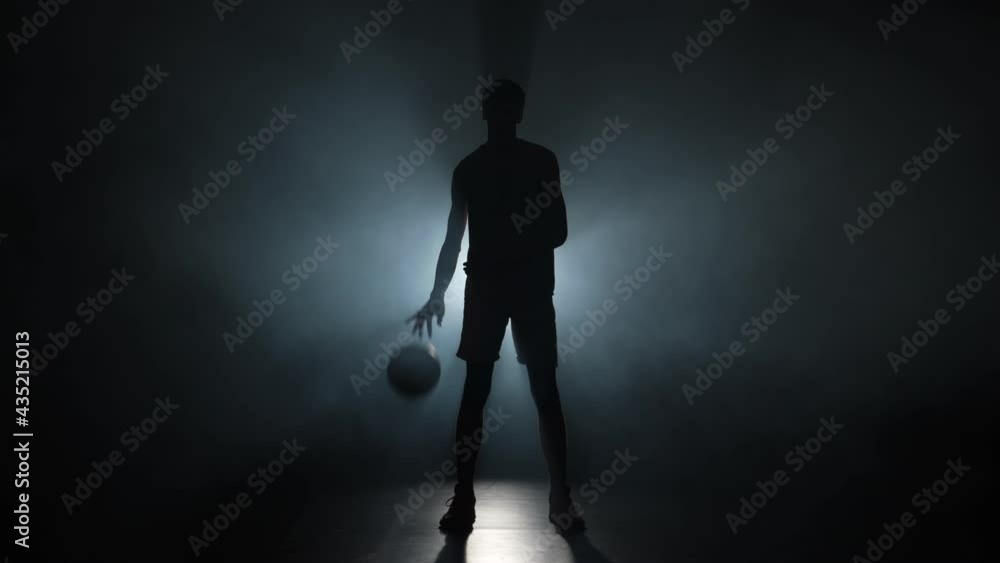 Silhouette of a young guy dribbling basketball on a dark studio background with smoke and backlight. Tall young sportsman in sports uniform hits ball on the floor. Slow motion ready 59.94fps. Silhouette of a young guy dribbling basketball on a dark studio background with smoke and backlight. Tall young sportsman in sports uniform hits ball on the floor. Slow motion ready 59.94fps.