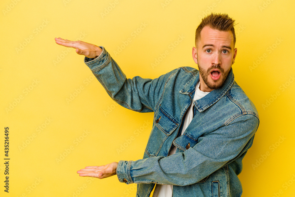 Young tattooed caucasian man isolated on yellow background shocked and amazed holding a copy space between hands. Young tattooed caucasian man isolated on yellow background shocked and amazed holding a copy space between hands.
