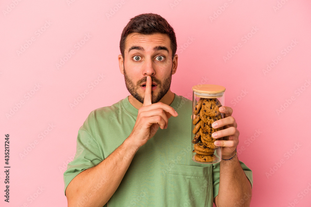 Young caucasian man holding cookies jar isolated on pink background keeping a secret or asking for silence. Young caucasian man holding cookies jar isolated on pink background keeping a secret or asking for silence.