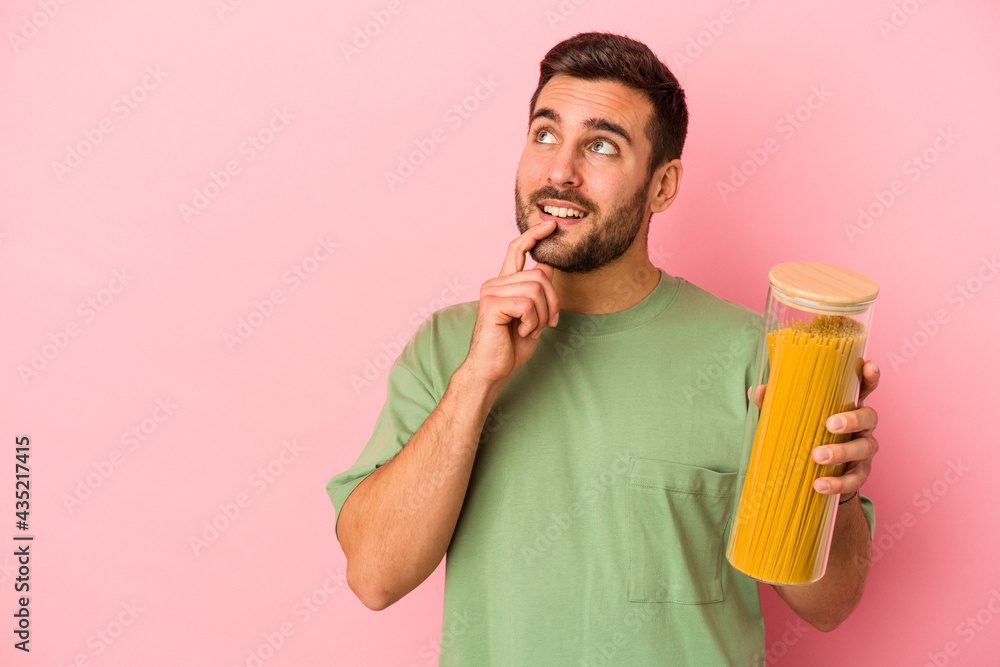 Fototapeta premium Young caucasian man holding pasta jar isolated on pink background relaxed thinking about something looking at a copy space.