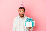 Young caucasian man celebrating his birthday isolated on pink background shrugs shoulders and open eyes confused.
