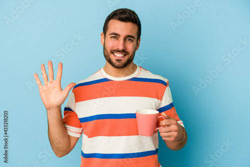 Young caucasian man holding a mug isolated on blue background smiling cheerful showing number five with fingers. © Asier