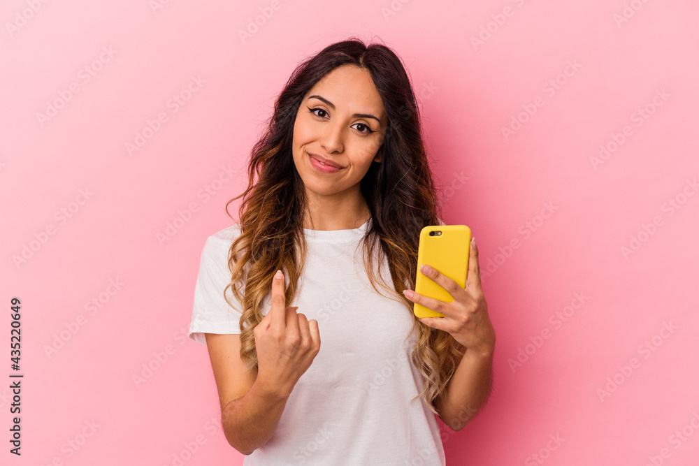 Young mexican woman holding a mobile phone isolated on pink background pointing with finger at you as if inviting come closer. Young mexican woman holding a mobile phone isolated on pink background pointing with finger at you as if inviting come closer.