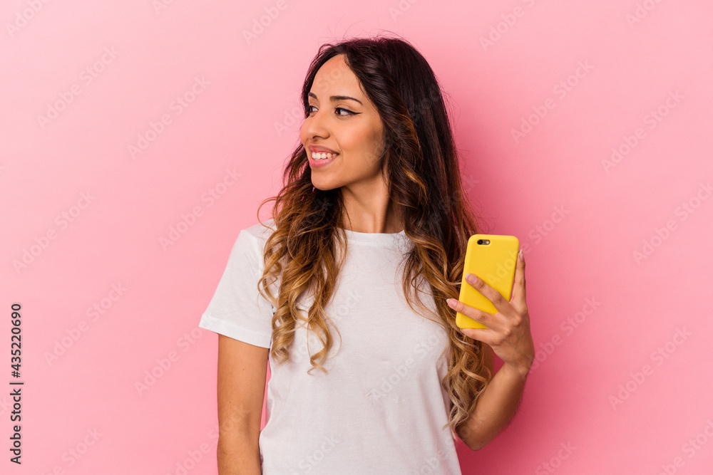 Young mexican woman holding a mobile phone isolated on pink background looks aside smiling, cheerful and pleasant. Young mexican woman holding a mobile phone isolated on pink background looks aside smiling, cheerful and pleasant.
