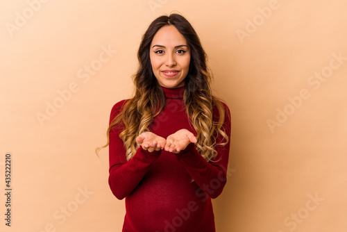 Young mexican pregnant woman isolated on beige background holding something with palms, offering to camera. © Asier