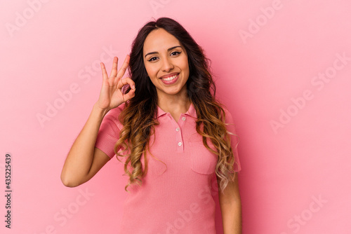 Young mexican woman isolated on pink background winks an eye and holds an okay gesture with hand.