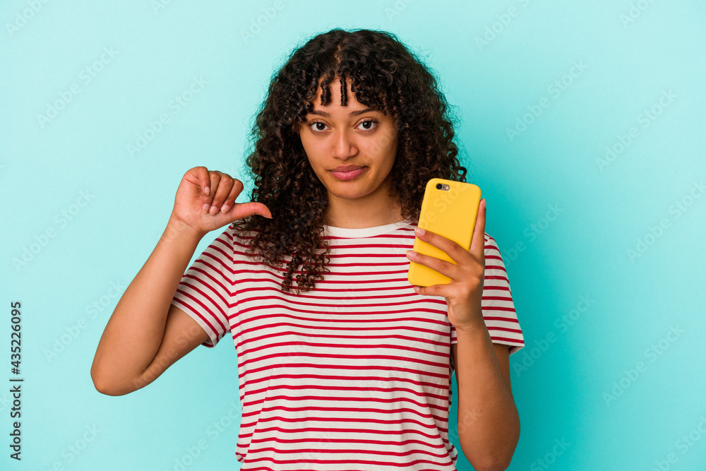 Young mixed race woman holding a mobile phone isolated on blue background showing a dislike gesture, thumbs down. Disagreement concept. Young mixed race woman holding a mobile phone isolated on blue background showing a dislike gesture, thumbs down. Disagreement concept.