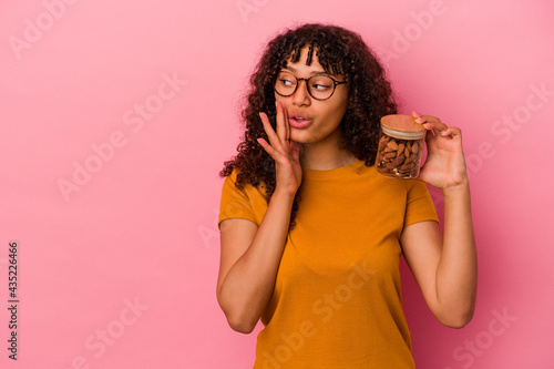 Young mixed race woman holding an almond jar isolated on pink background is saying a secret hot braking news and looking aside