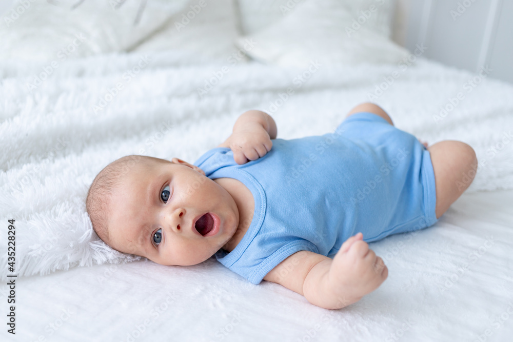 yawning baby boy three months old in a blue bodysuit on a white bed at home goes to bed yawning baby boy three months old in a blue bodysuit on a white bed at home goes to bed