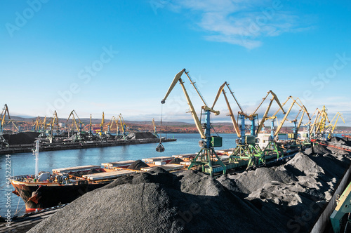 Heaps of coal in the Murmansk Commercial Sea Port. Loading of coal by buckets (grabs) of portal cranes into the holds of a dry cargo ship in the seaport
