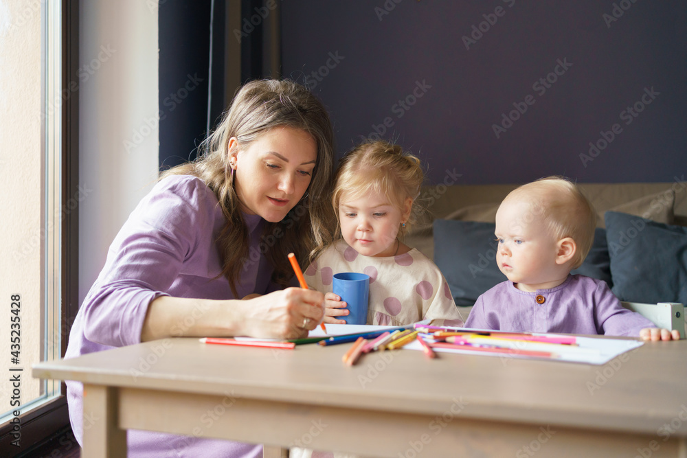 Young loving mother drawing with colored pencils with two adorable little kids at home. Happy mom teaching small daughter and baby boy son how to draw. Motherhood and mothers day concept Young loving mother drawing with colored pencils with two adorable little kids at home. Happy mom teaching small daughter and baby boy son how to draw. Motherhood and mothers day concept