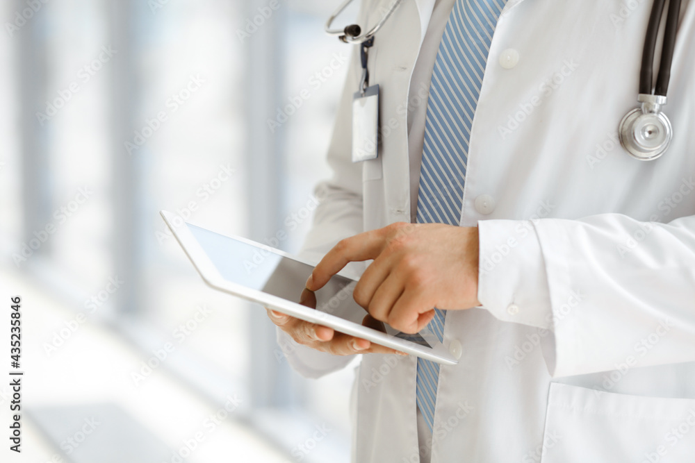 The unknown male doctor is using a tablet computer while standing in a modern clinic, close-up. Medicine concept