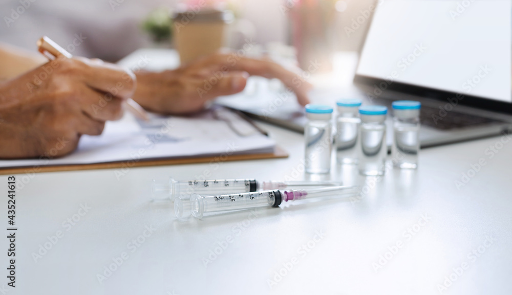 Focus on the syringe on table and blurred hand of doctor working with laptop and searching data of corona virus Focus on the syringe on table and blurred hand of doctor working with laptop and searching data of corona virus