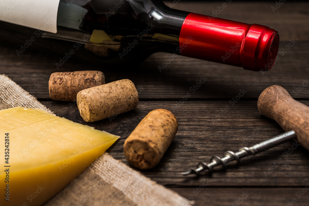 Bottle of red wine with a piece of parmesan lying on a piece of canvas and corkscrew with corks lying on an old wooden table. Close up view, focus on the bottle of red wine
