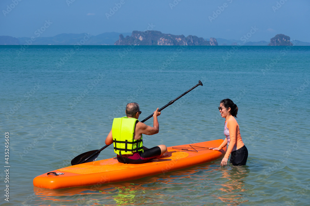 asian senior father playing standing up paddleboard or SUP with young daughter at blue sea on summer vacation. family together concept