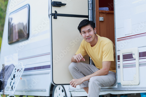 Young adult Asian man sitting in camper van or RV parking at camping location. Attractive Asian man relaxed in the motor home by the nature on summer.