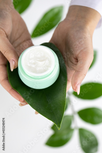 Close-up hands hold cosmetic cream jar on fresh green leaf on white herbal background. Skinimalism, skincare routines, sustainable packaging, eco-conscious, beauty, plant-based cosmetology concept