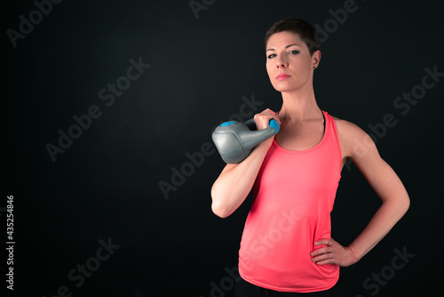 Attractive sporty woman with short hair ready to press kettle bell with her right hand. Woman athlete in indoor sportswear agains dark backround. Left hand on the hip