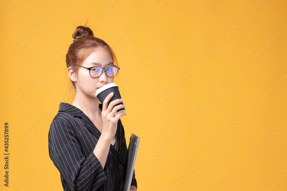 Obraz premium Portrait of a beautiful young Asian woman student wear glasses holding laptop and drinking coffee isolated on yellow background. Beautiful young girl enjoys coffee in the morning.