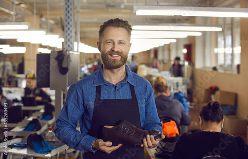 Portrait of footwear factory worker with brand new product. Happy young man standing in workshop, holding new leather boots, looking at camera and smiling. Shoe manufacturing industry concept