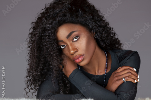 Portrait of a sensual young black woman with long curly black hair, beautiful makeup sitting by herself on fur in a studio with a grey background wearing a black long sleeve shirt and jewelry.