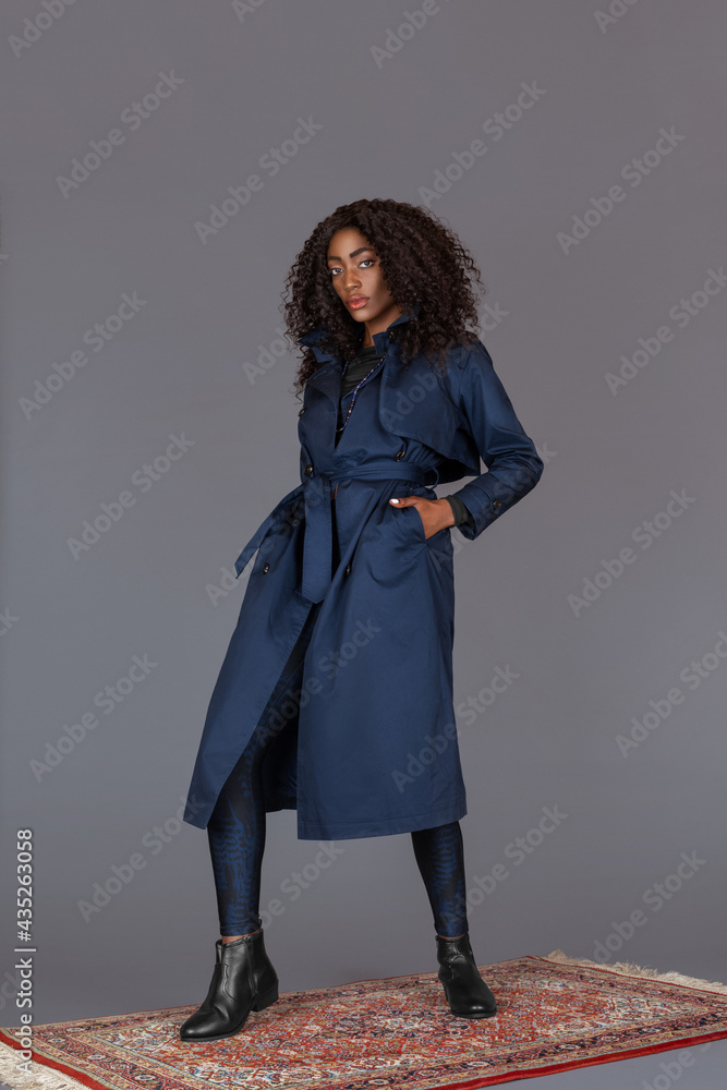 Portrait of a sensual black woman with long curly black hair, beautiful makeup posing by herself on a carpet in a studio with a grey background wearing a blue trench coat and leggings with black boots Portrait of a sensual black woman with long curly black hair, beautiful makeup posing by herself on a carpet in a studio with a grey background wearing a blue trench coat and leggings with black boots