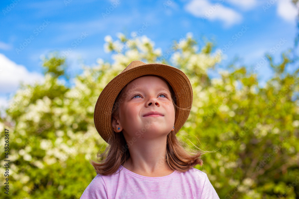 A beautiful little girl in a hat looks at the sky. A child in a blooming garden.