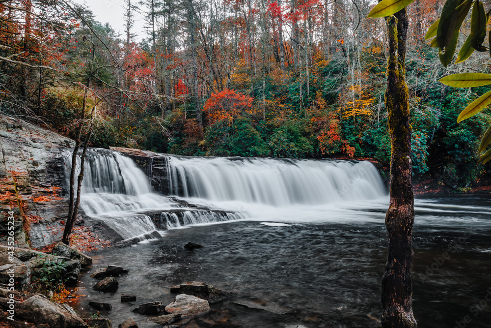 Fototapeta premium Hooker Falls, a waterfall of the Little River surrounded by colorful fall foliage on a gloomy, cloudy day in DuPont State Recreational Forest, Hendersonville, North Carolina, USA. Long exposure.