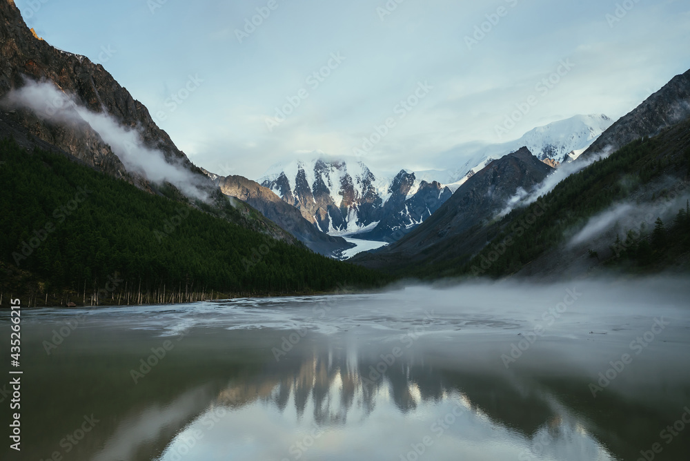 Atmospheric alpine landscape with snowy mountains in golden sunlight reflected on mirror mountain lake in fog among low clouds. Scenic highland scenery with low clouds on rocks and green mirror lake.