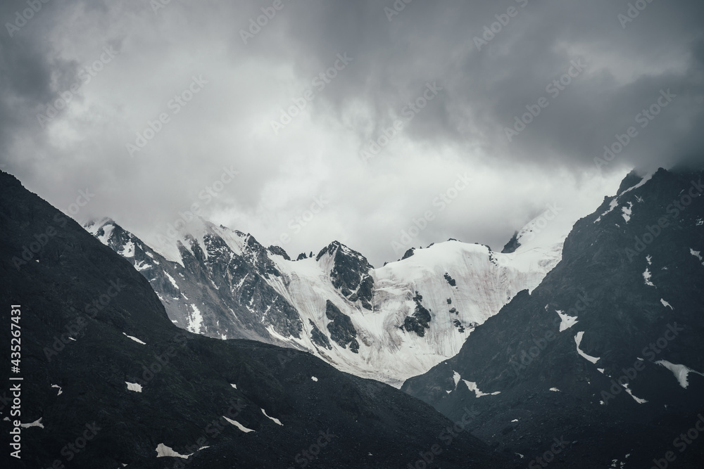 Dark atmospheric mountain landscape with glacier on black rocks in lead gray cloudy sky. Snowy mountains in gray low clouds in rainy weather. Gloomy mountain landscape with rocky mountains with snow.