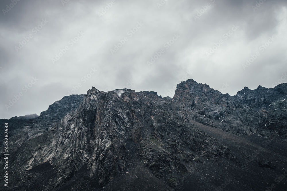 Dark atmospheric landscape with rocky mountain wall under gray cloudy sky. Rocky pinnacle in lead gray cloudy sky. Dark mountain peak in overcast weather. Gloomy minimalist scenery with high mountains