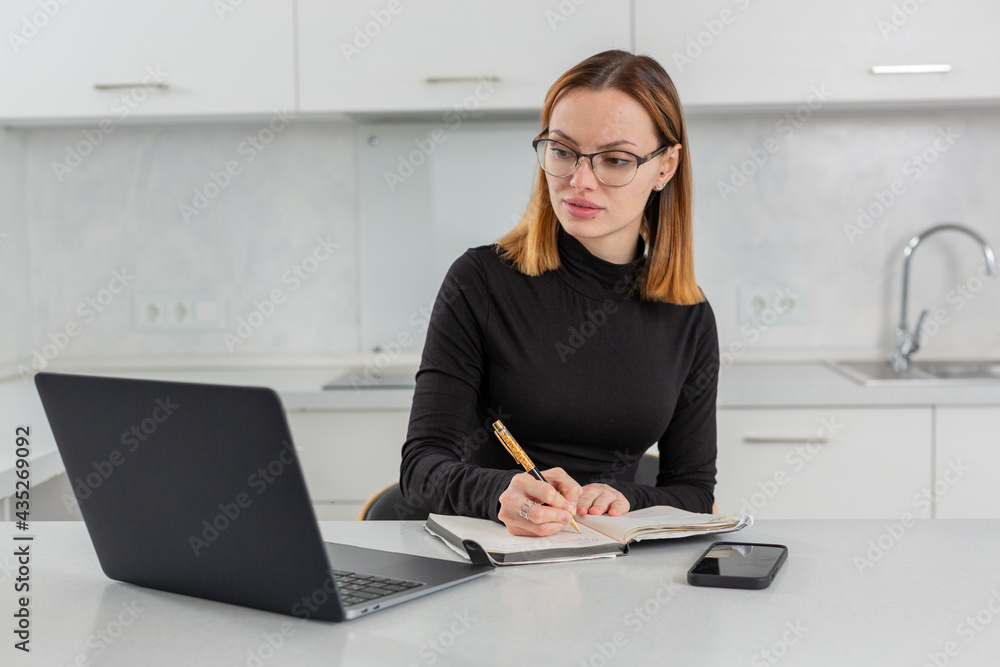 a girl with glasses makes notes in a notebook, working for a laptop at home in the kitchen, looking at the screen of the monitor. High quality photo a girl with glasses makes notes in a notebook, working for a laptop at home in the kitchen, looking at the screen of the monitor. High quality photo