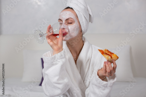 portrait of a girl in a white robe and a moisturizing face mask, who drinks red wine from a glass, eats pizza while sitting in her hotel room. High quality photo